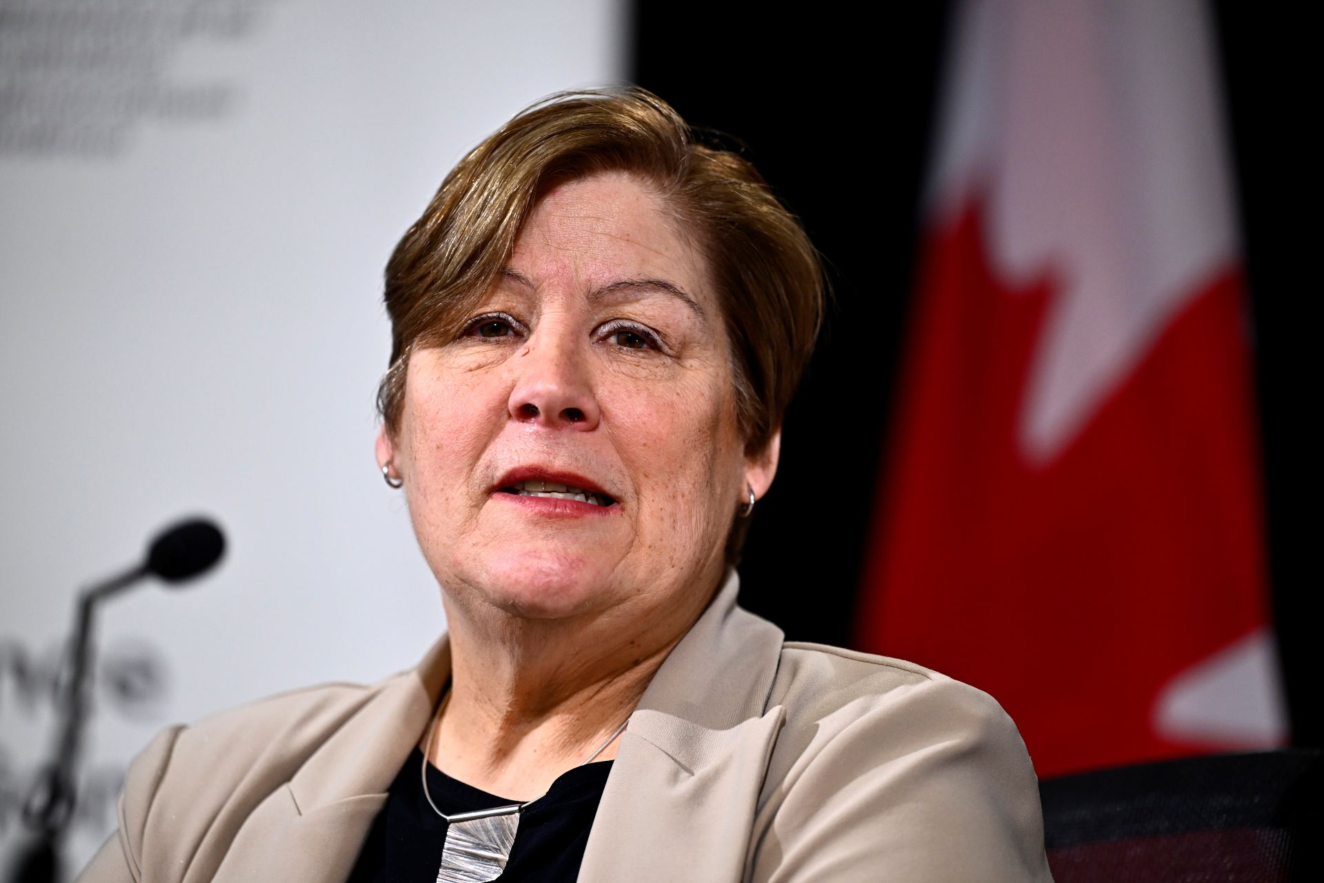 A closeup of Hogue speaking in front of a microphone with a Canadian flag in the backdrop. 