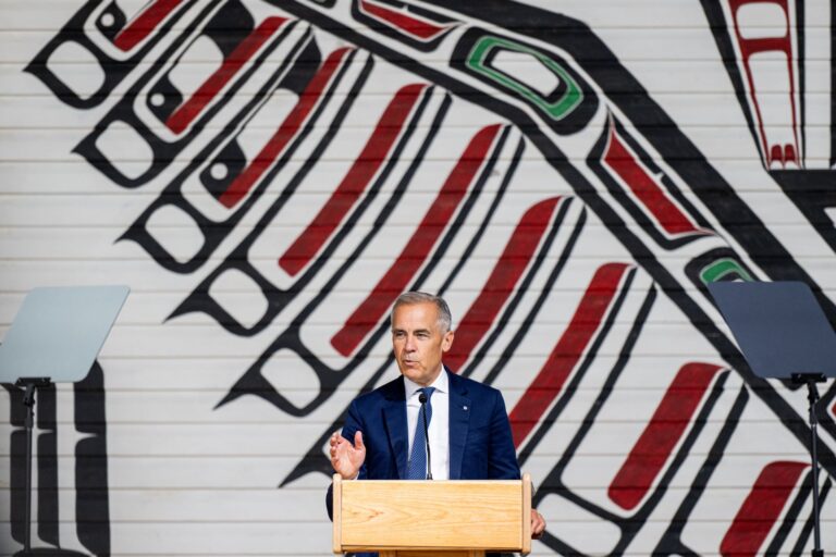 Carney stands in front of a painting that is twice as tall as he is. It depicts a bird’s wing in black lines with red and green accents on a white wood surface.