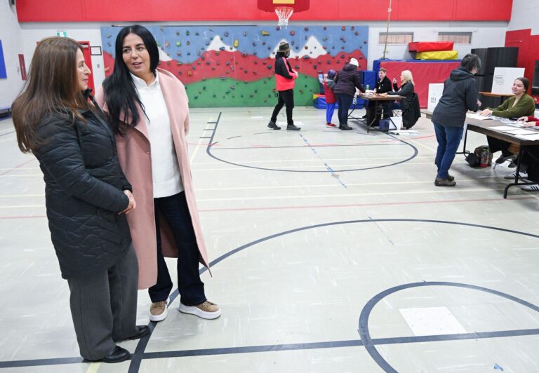 People voting at two polling stations in a school gymnasium while the candidate and her mother wait.