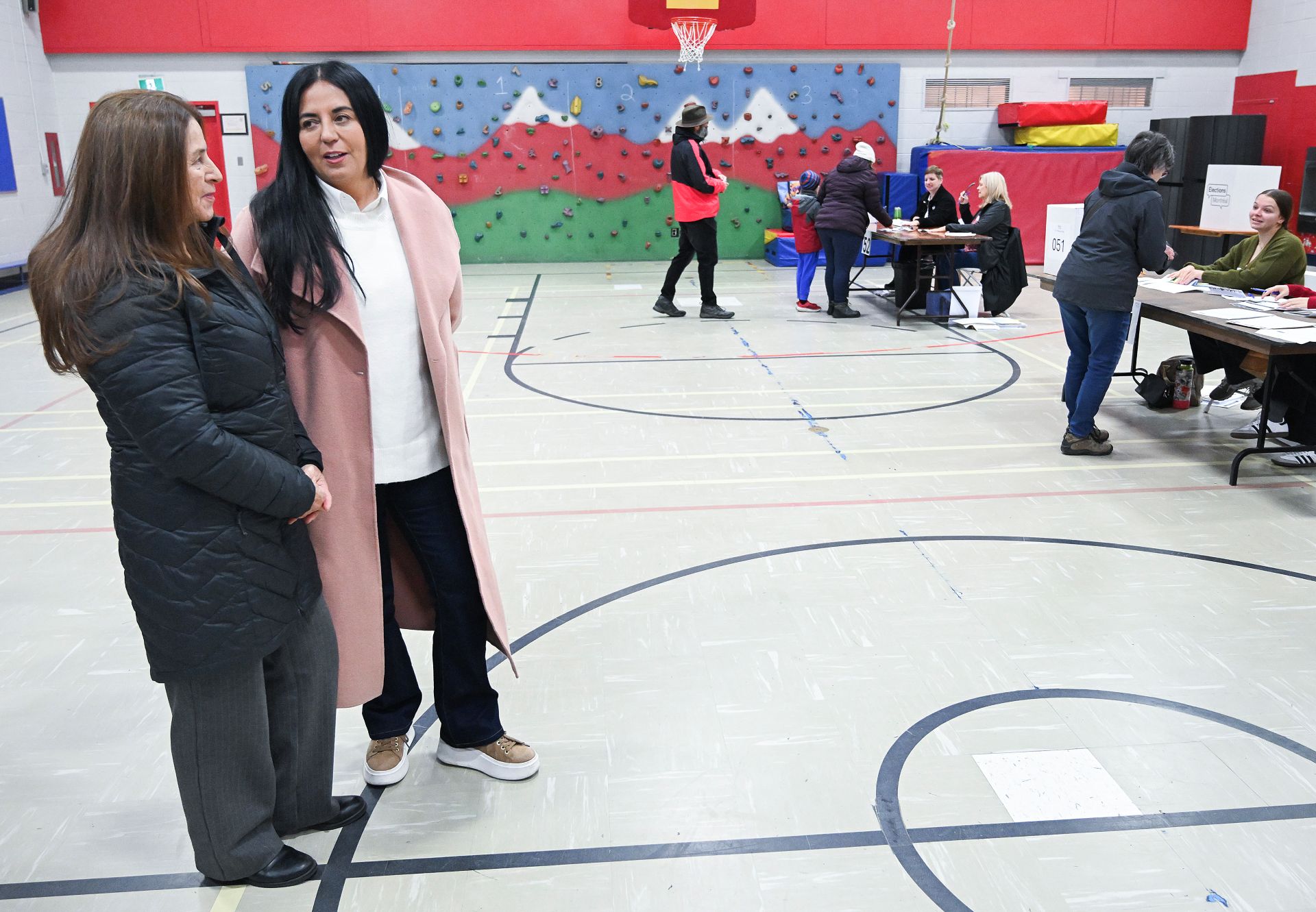 People voting at two polling stations in a school gymnasium while the candidate and her mother wait.