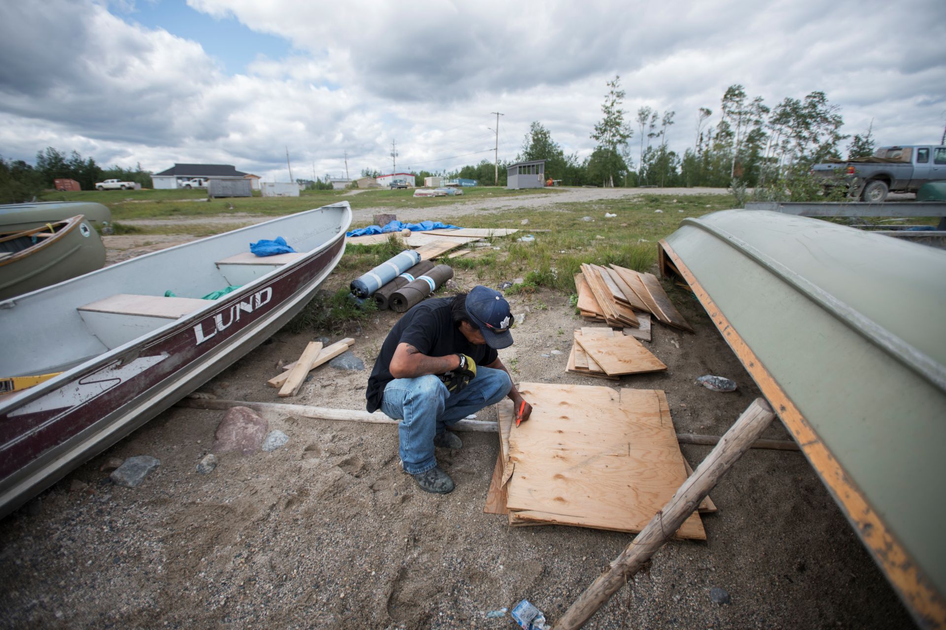 A man squats on the sandy shore of a lake, working with sheets of plywood. He is surrounded by metal motor boats sitting on the shore. 