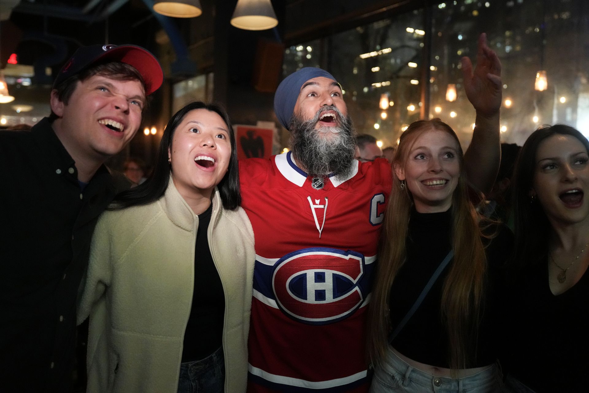 Jagmeet Singh wide-eyed and enthusiastic watching a screen with his arms over some supporters.