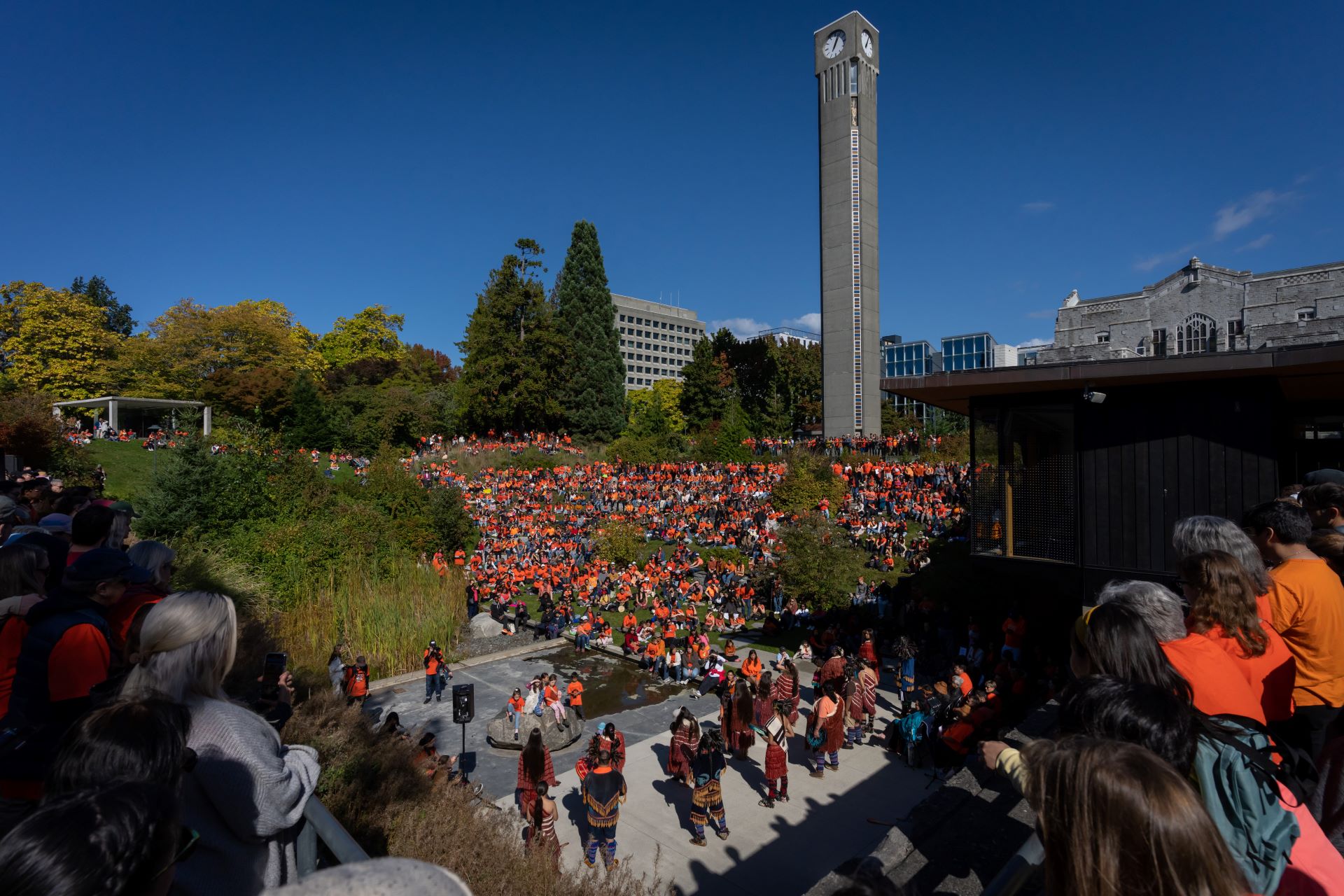 ALT TEXT: Hundreds of people in orange shirts are standing and seated in an area bordered by trees and buildings.