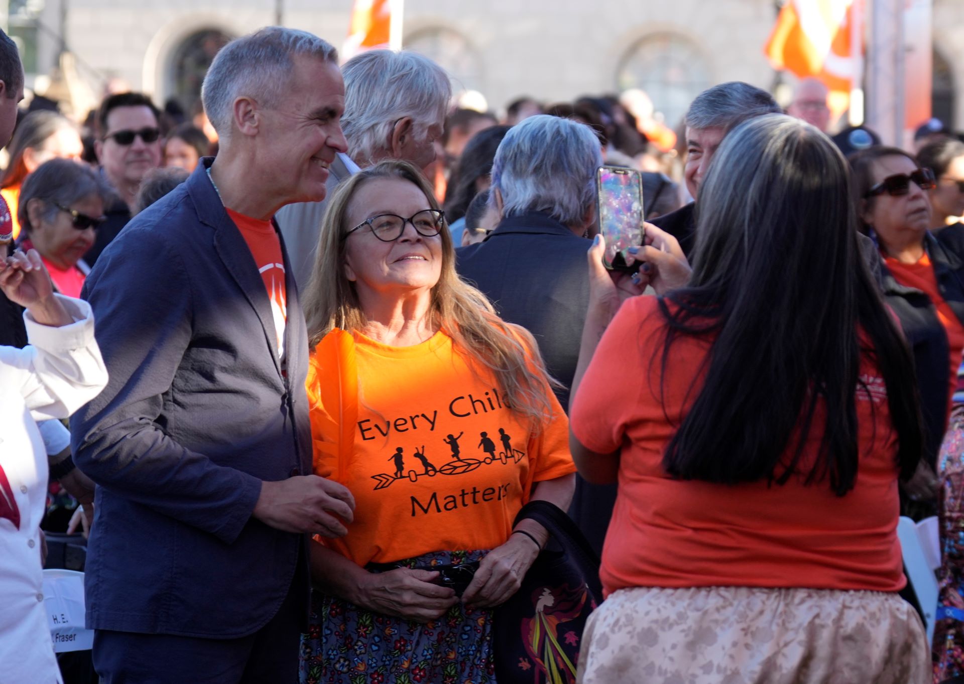 Carney is in a casual black jacket with an orange shirt underneath. A woman in an orange shirt is using her phone to take a picture of him and the woman among a crowd of people.