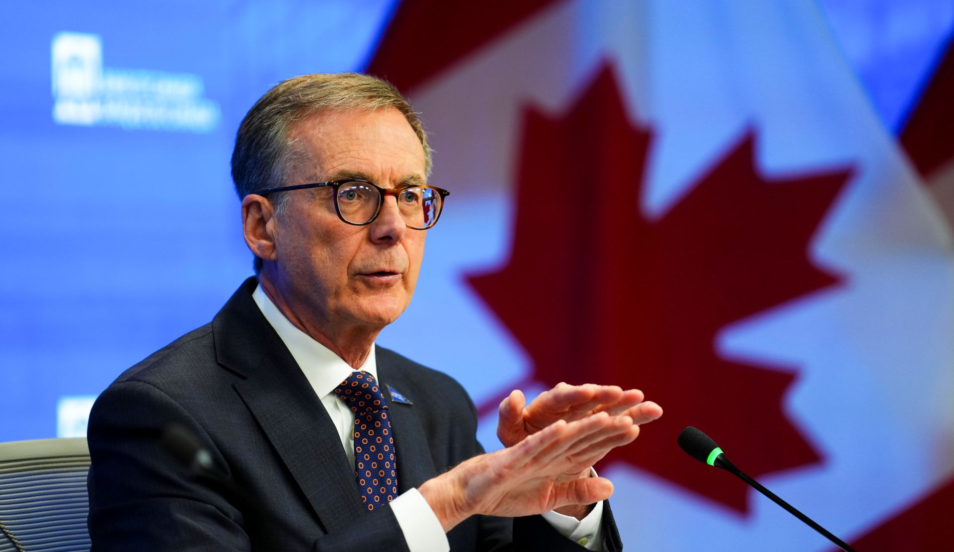 Tiff macklem gestures with his hands while speaking at a microphone with a Canadian flag in the background.