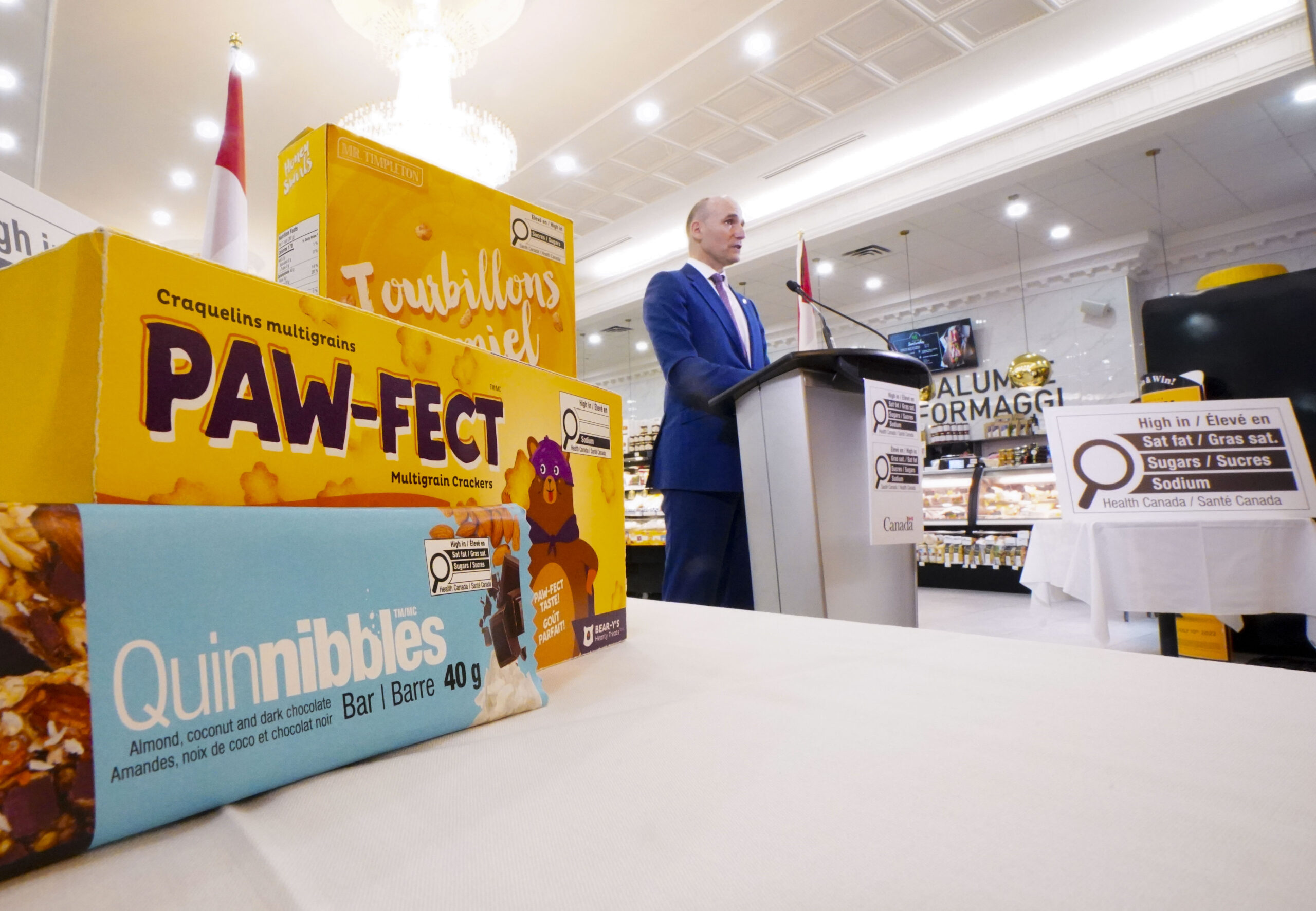 Un homme s’exprime au pupitre lors d’une annonce sur l’étiquetage alimentaire, avec des produits emballés au premier plan