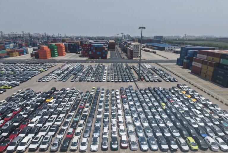 Hundreds of cars parked in a lot with containers and cranes in the background.