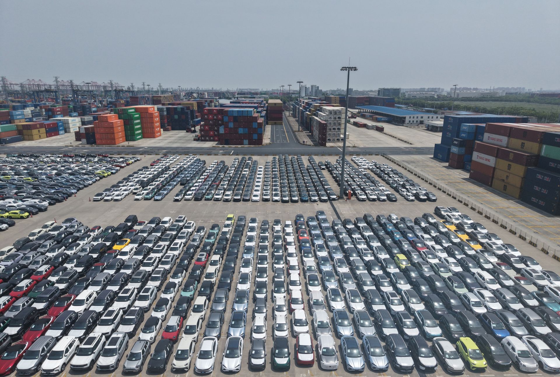 Hundreds of cars parked in a lot with containers and cranes in the background.