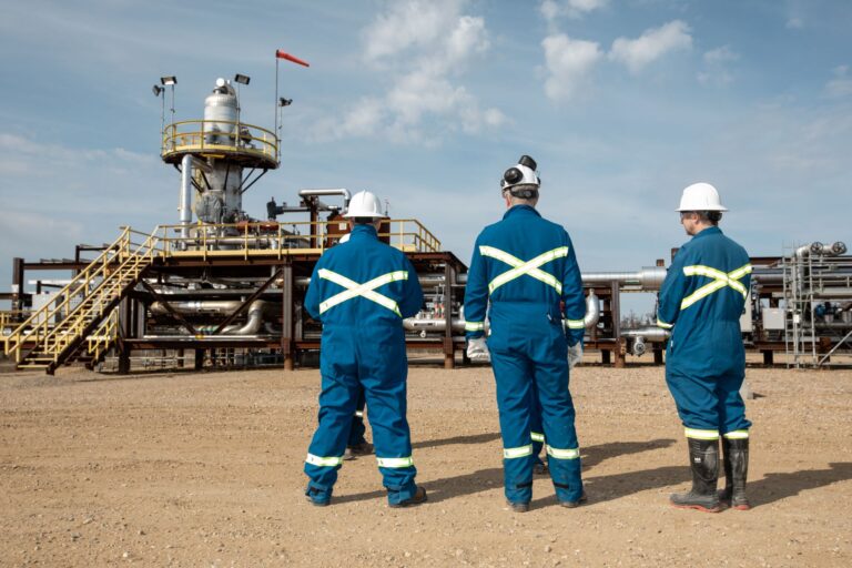 Three men in blue coveralls stand side by side looking at machinery in the distance.