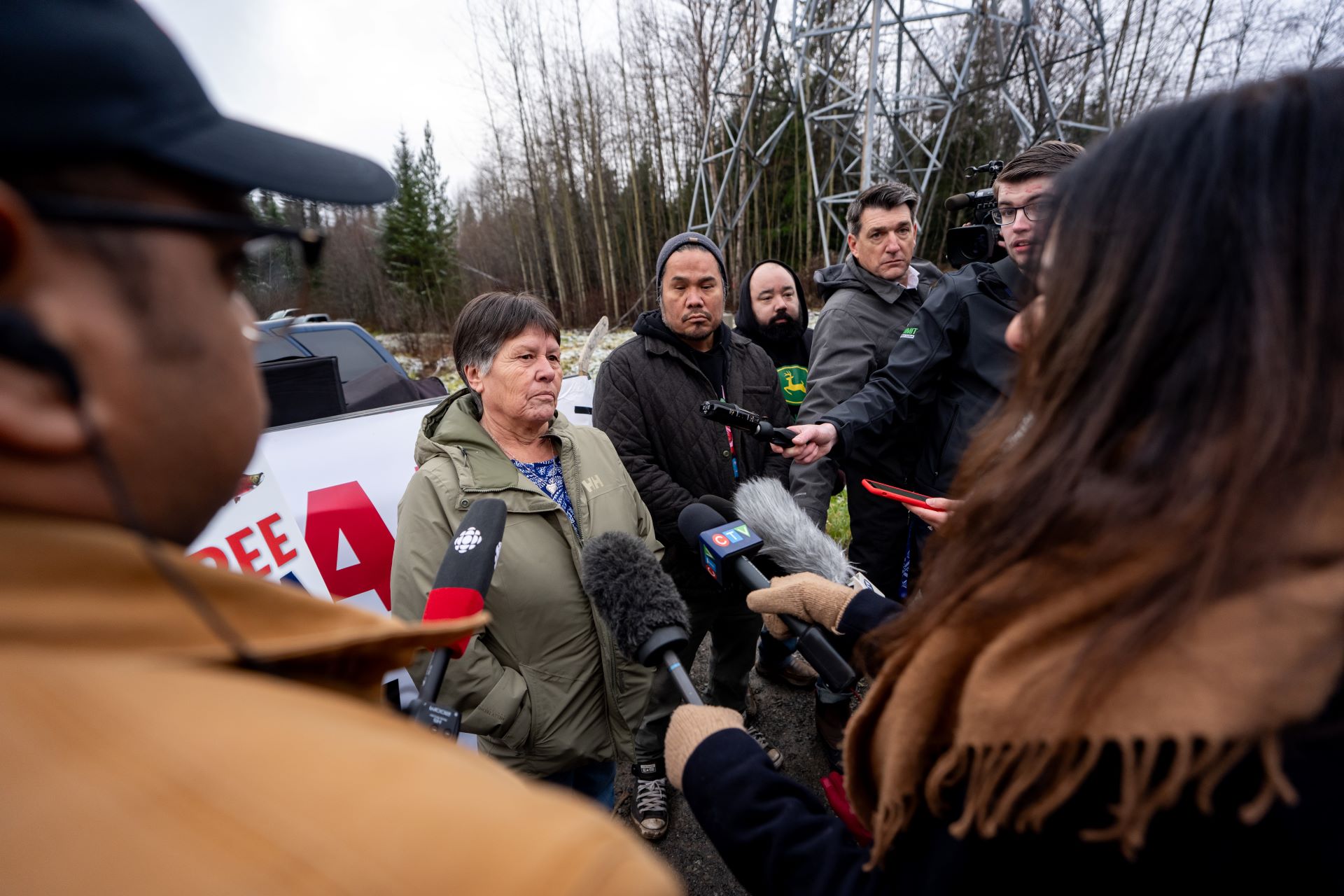 A small group of reporters surrounds the chief with microphones outstretched toward her. They are standing in a clearing of a wooded area next to a metal electricity tower.