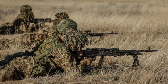 Four soldiers in camouflage laying in dead grass firing weapons.