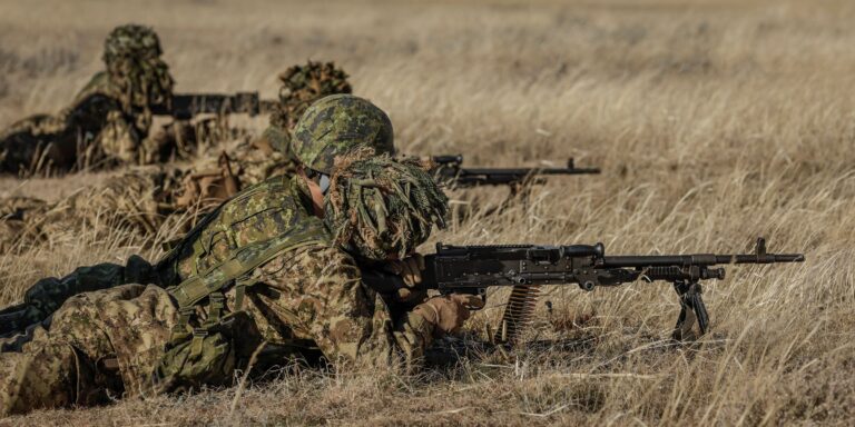 Four soldiers in camouflage laying in dead grass firing weapons.