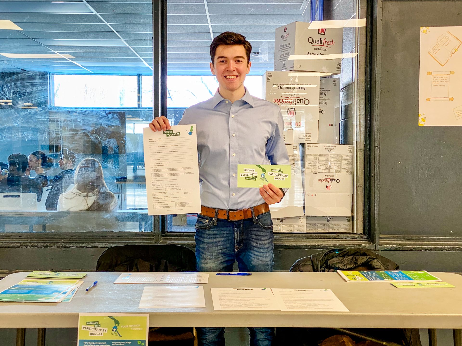 Lecchino stands behind a long table, holding up a piece of paper in each hand.