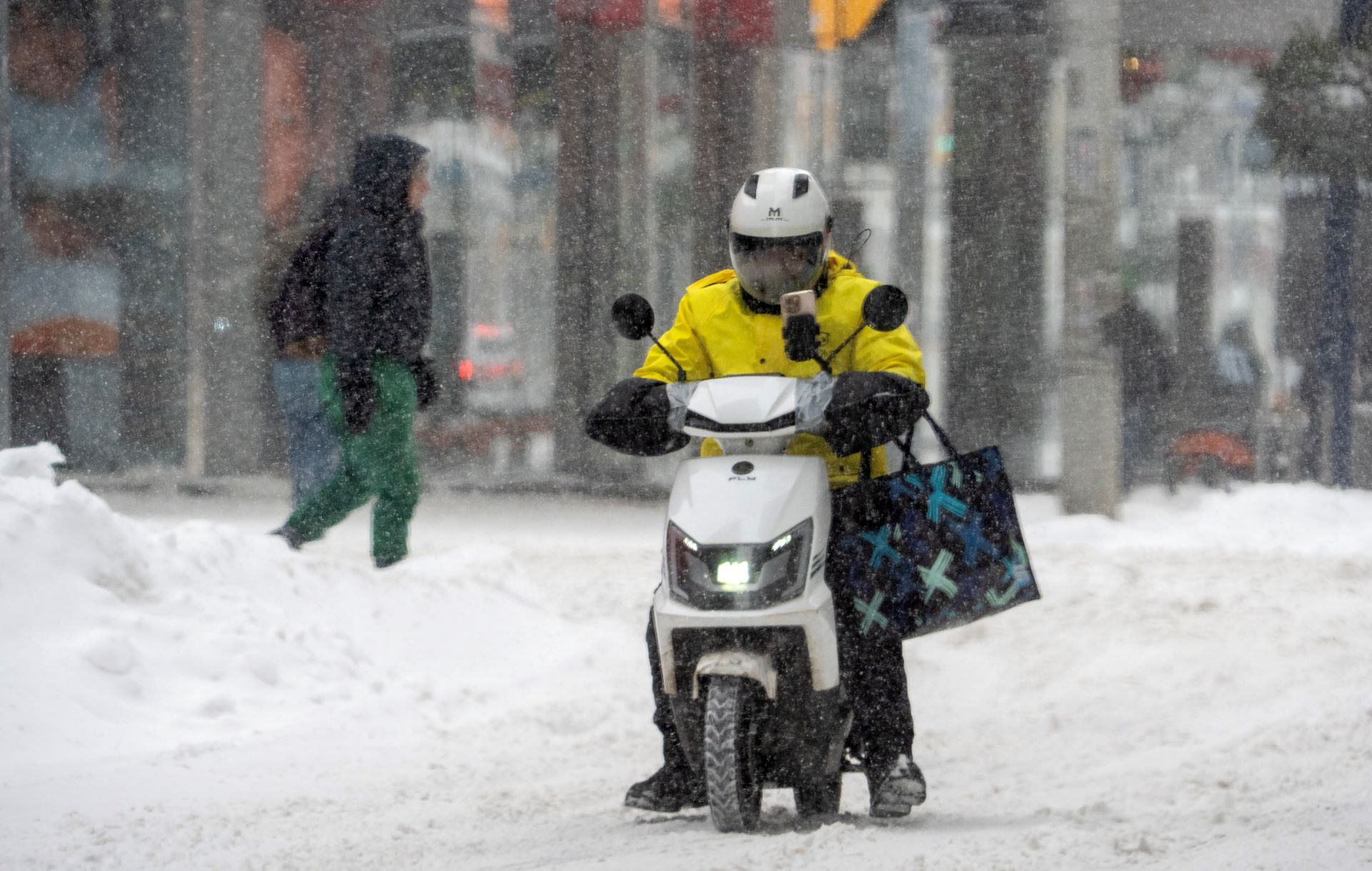 The driver is in a heavy yellow jacket and white helmet, with a black shopping bag hanging from one arm. He’s stopped or going slow on a road covered in snow.