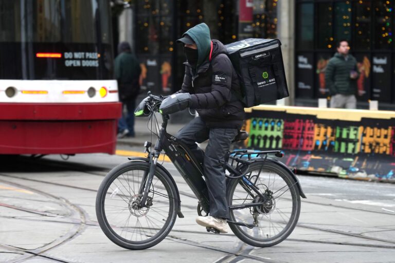 A man on an e-bike crosses an intersection with a boxy insulated food bag strapped to his back. He’s wearing bulky mitts, snow pants, and a cover for his nose and mouth under a ball cap and hoodie with a puffer jacket.