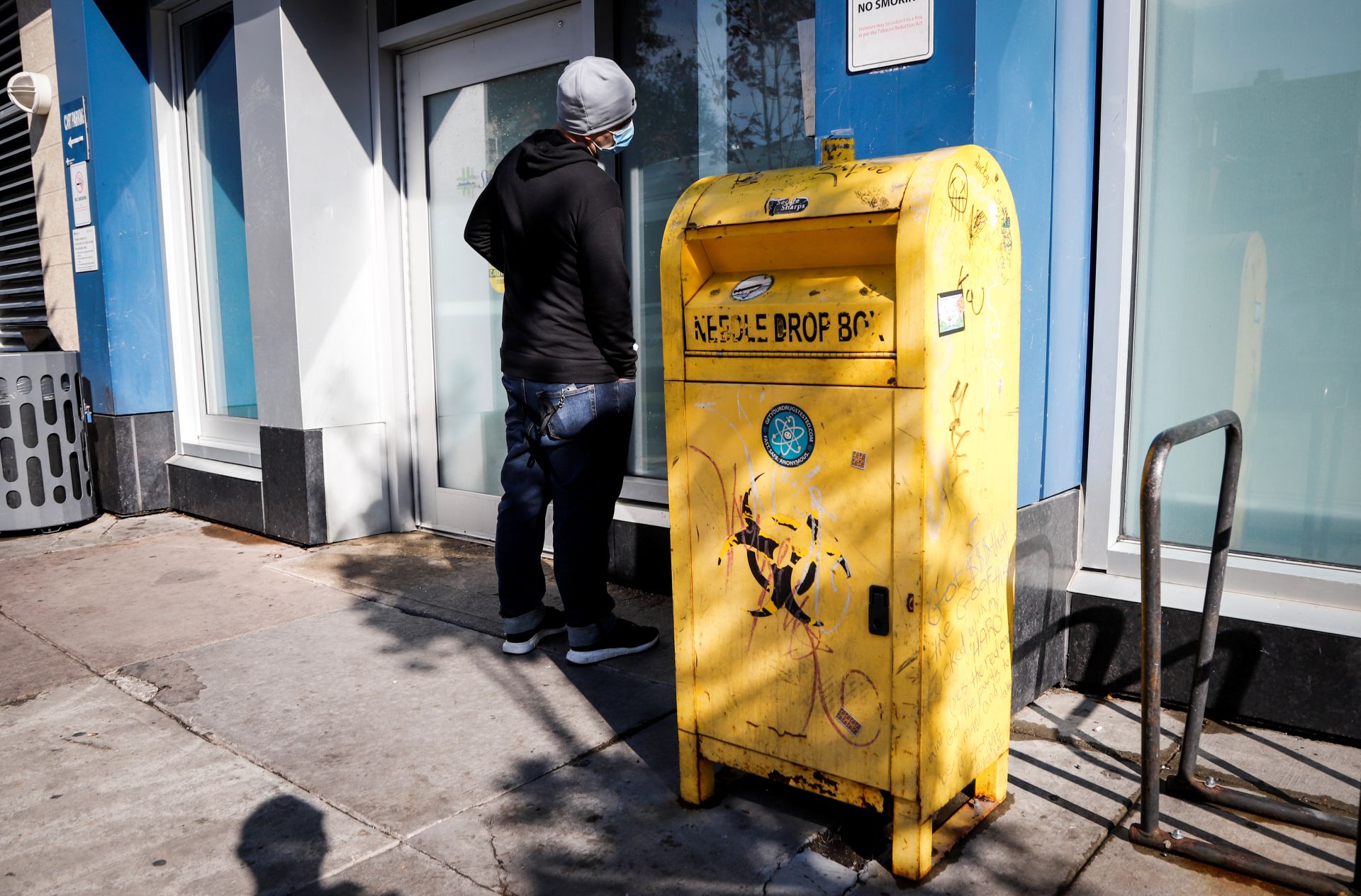 A man stands next to a yellow needle drop box that resembles the shape of a mailbox as he looks through a window.