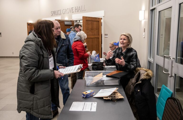 A half dozen people line up in front of a table as a woman explains something.