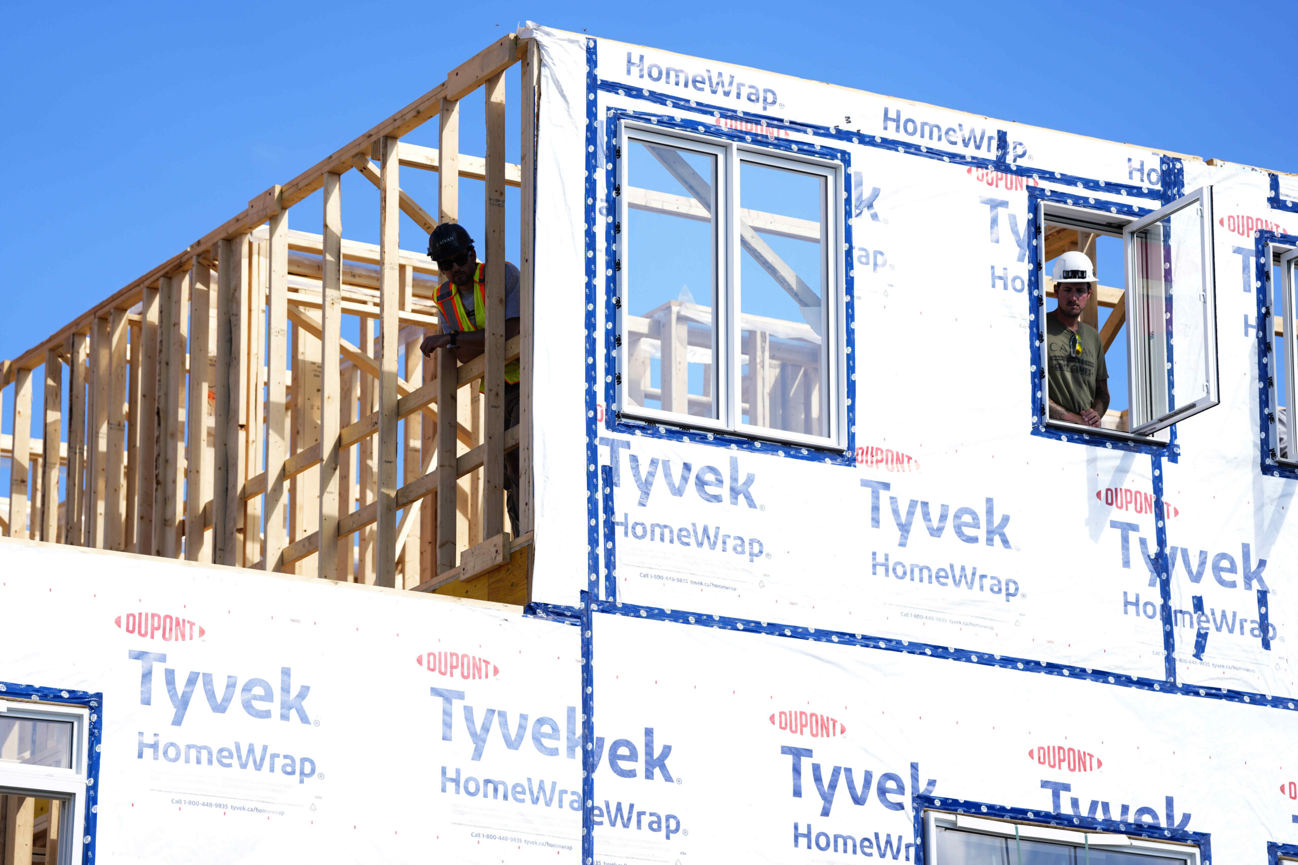 Des ouvriers travaillent sur la structure en bois d’une maison modulaire en construction, partiellement recouverte d’un pare-intempéries Tyvek, sous un ciel bleu.