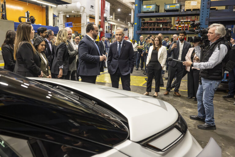 Le premier ministre du Canada se tient dans une usine de pièces automobiles, entouré de représentants de l’industrie, de travailleurs et de journalistes, devant un véhicule.
