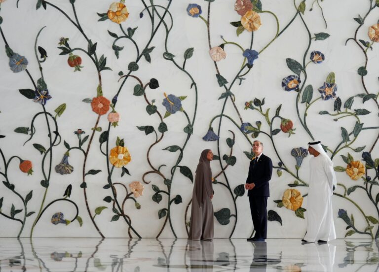 The three of them are standing in a room with at least double-height ceilings. Behind them, the wall is covered in an artwork consisting of intertwined vines with flowers and leaves.