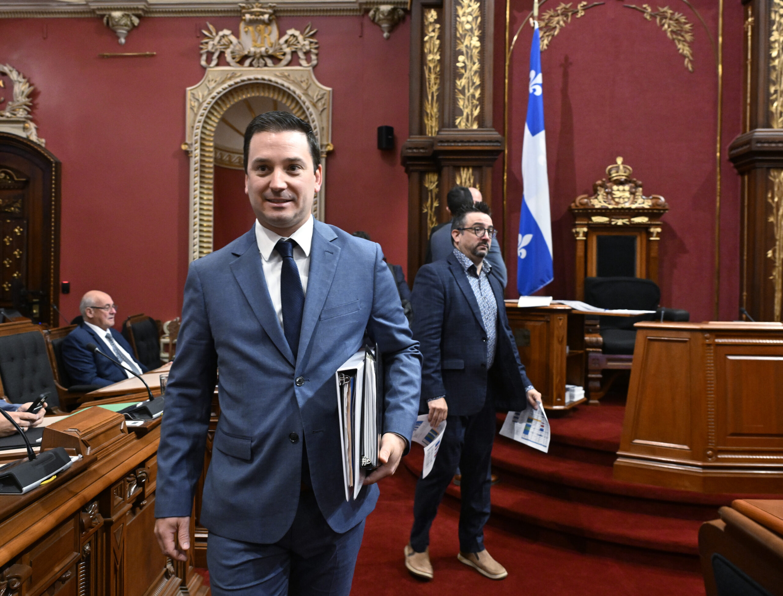 Scène à l’Assemblée nationale du Québec. Au premier plan, un homme en costume bleu clair et cravate sombre marche vers l’avant avec un porte-documents et des papiers sous le bras.