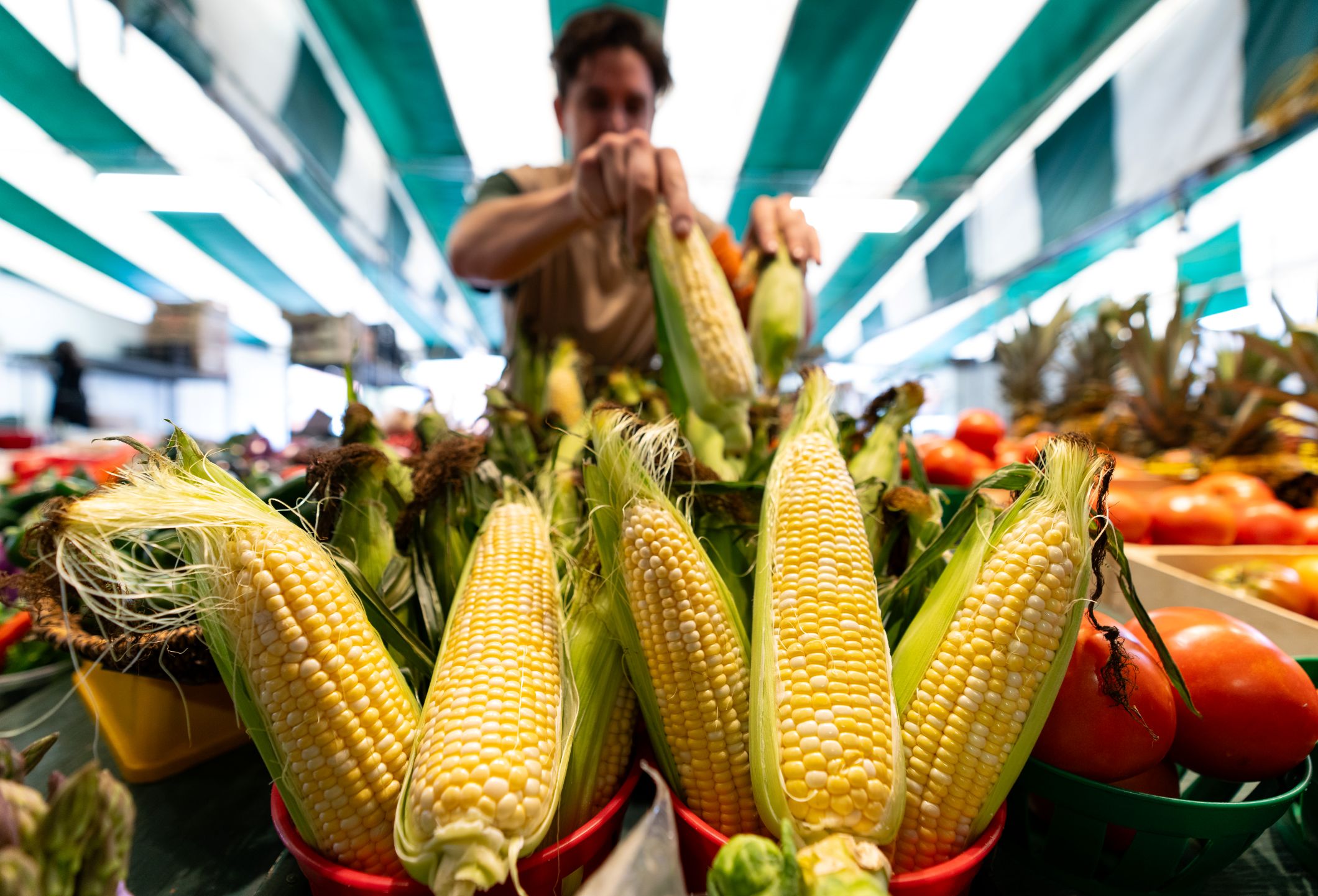 Corn on the cob in closeup, with other produce and a store worker in the blurry background.