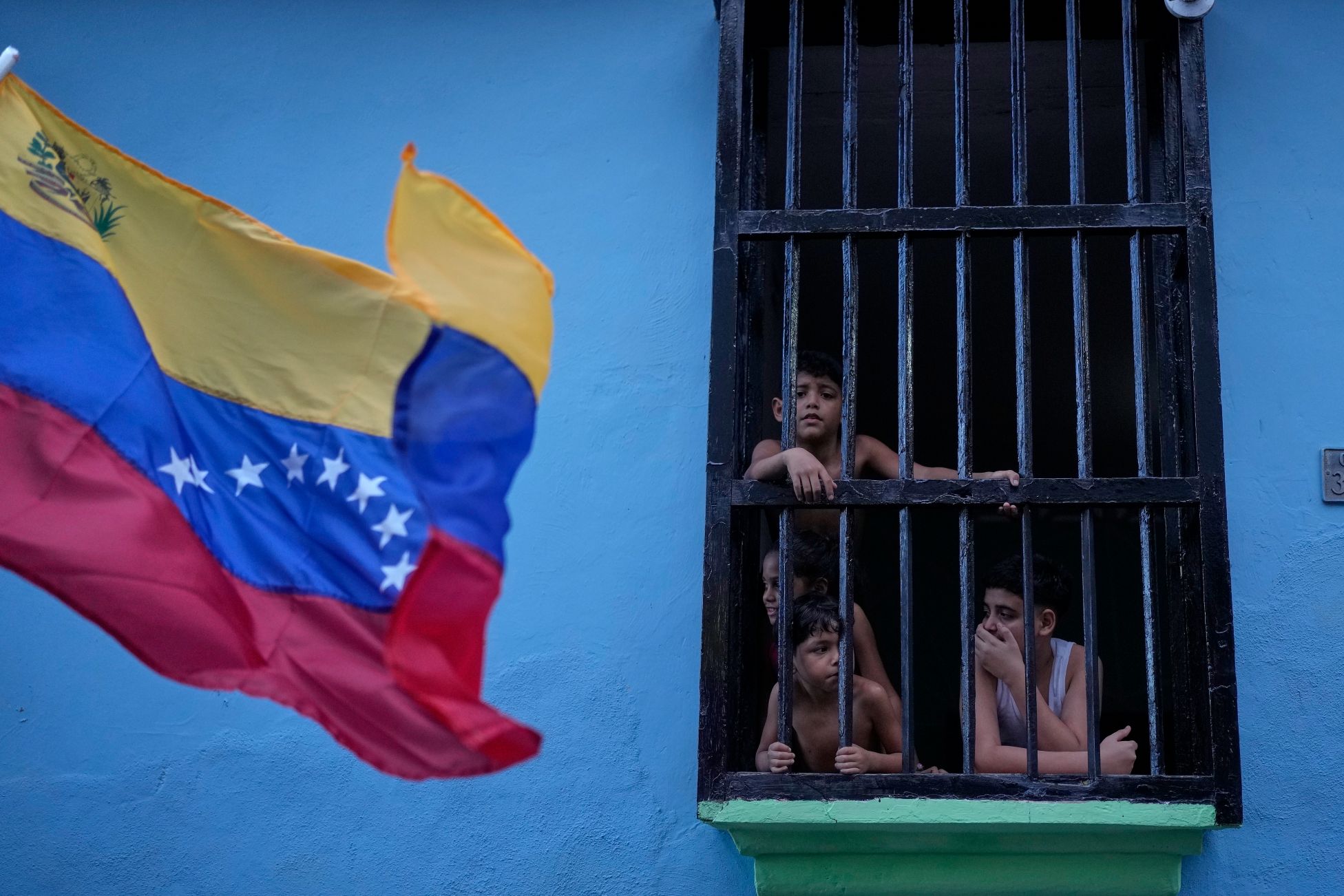 Three children look out from a window with bars on it as a Venezuelan flag passs by.