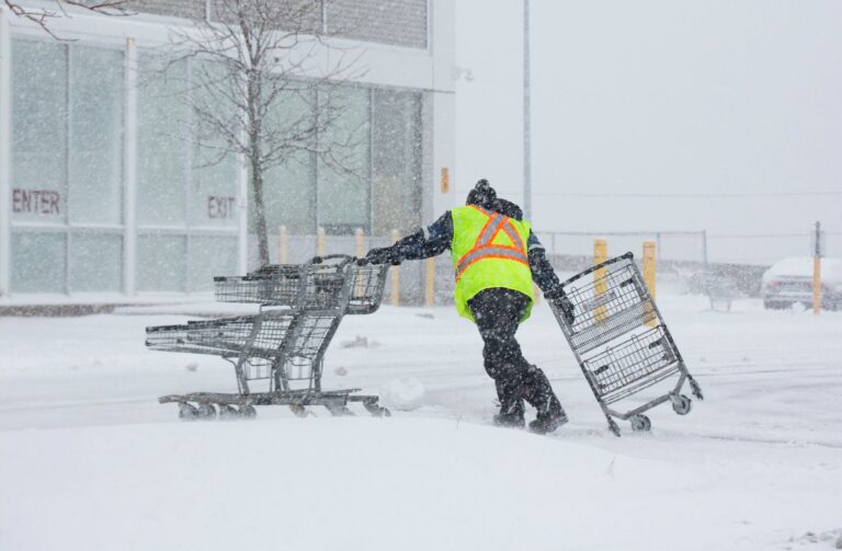 A worker in a yellow safety vest wrangles three shopping carts in a driving snowstorm.
