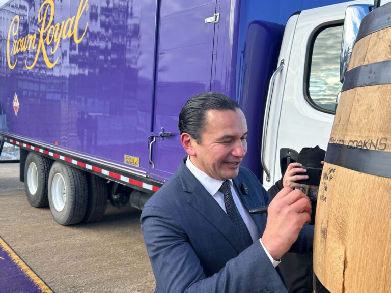 Wab Kinew signs a barrel with a truck with the purple Crown Royal sign on its side in the background.