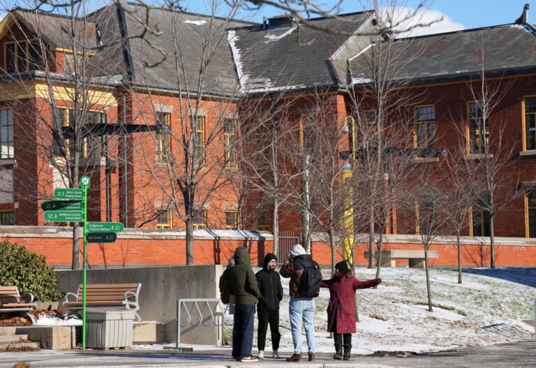 A group of four students bundled up in the cold standing in front of a nondescript brick building.