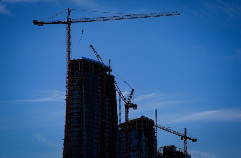 Three cranes working on three towers are in darkened silhouette against a dark blue sky.