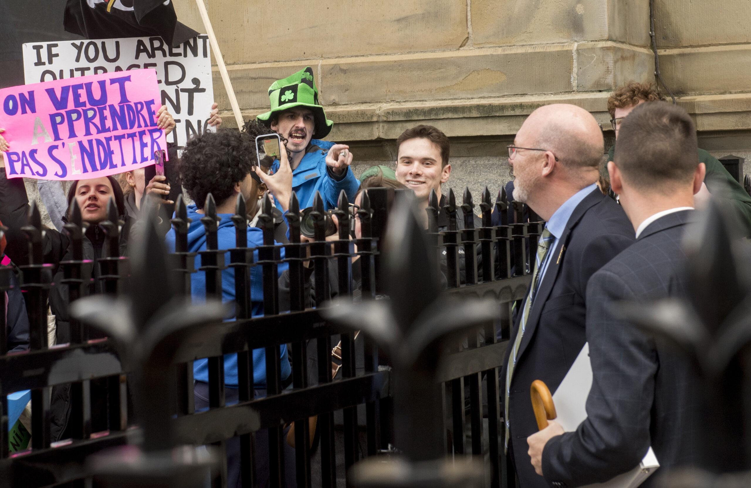 Un groupe d’étudiants manifeste derrière une clôture en fer devant un bâtiment en pierre, tenant des pancartes — dont l’une dit « On veut apprendre, pas s’endetter » — et scandant des slogans. Certains filment la scène avec leur téléphone. De l’autre côté de la clôture, un homme en complet, accompagné d’un autre, leur fait face en tenant des documents et un parapluie. L’ambiance est tendue et engagée.
