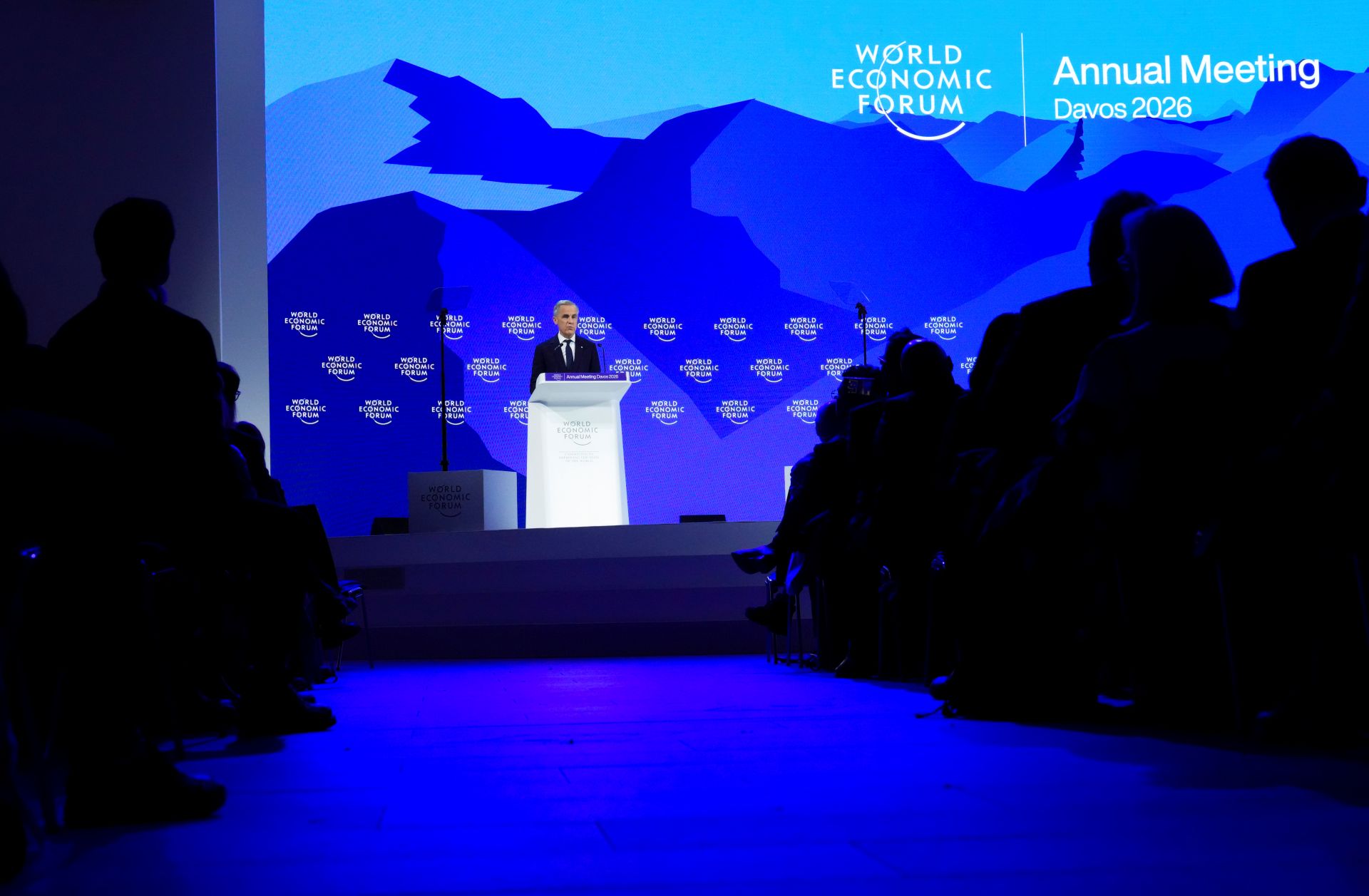 Carney is at the podium. The backdrop is a blue digital illustration of mountains.