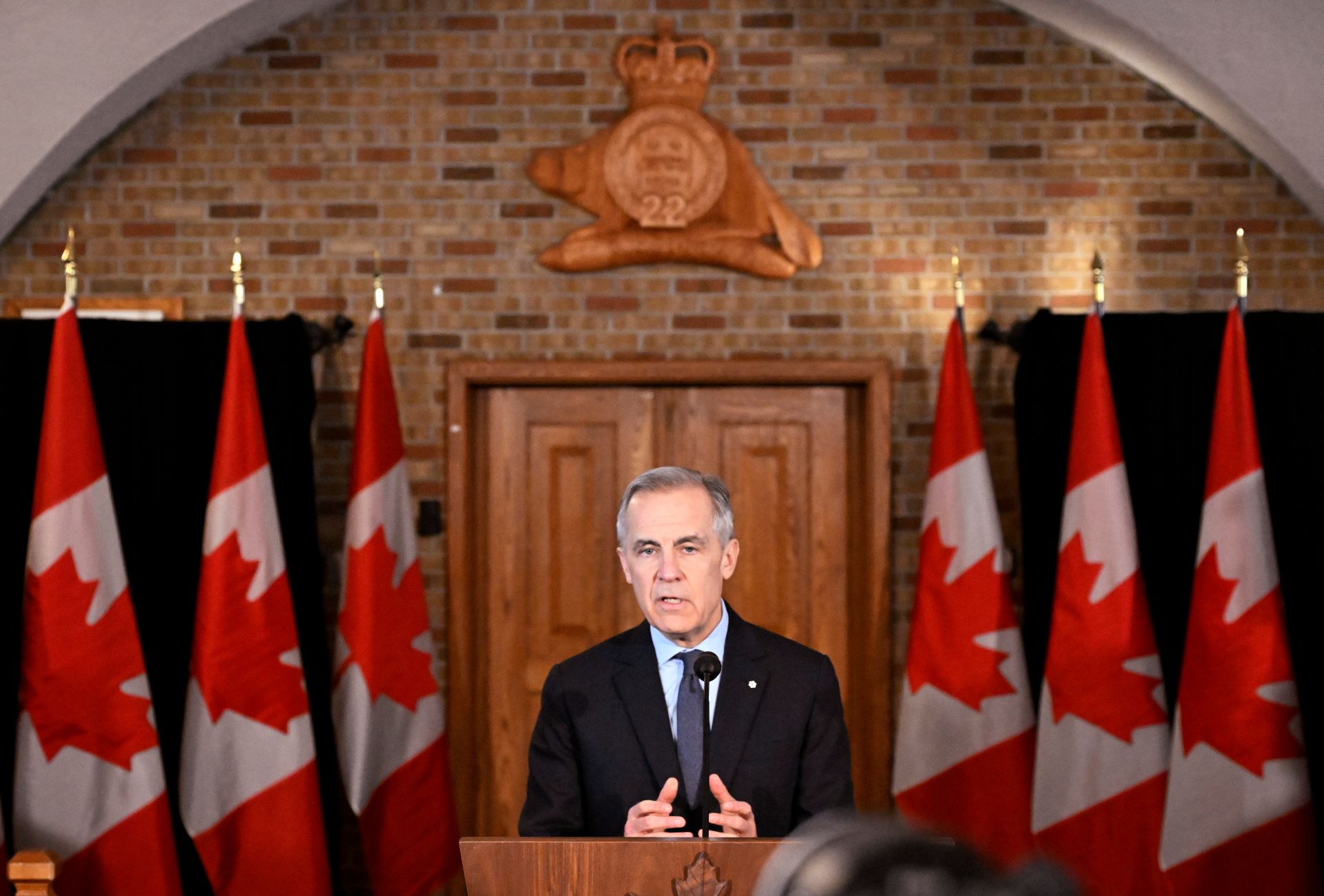 Mark Carney at a podium in front of wooden doors at the Citadelle, with the Royal 22e regiment emblem featuring 22 and a beaver above the door. There are also six Canadian flags. 