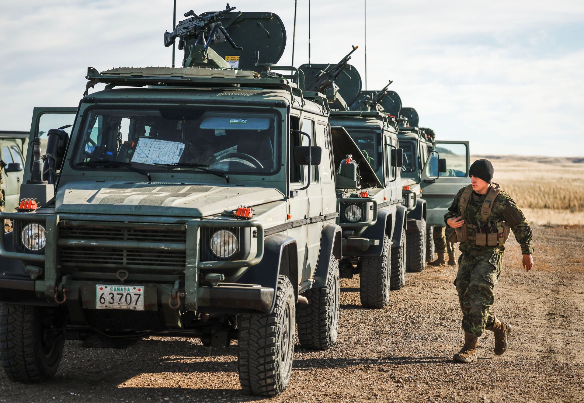 A line of G-Wagon SUV-style vehicles, but in army green with many antennae. A soldier approaches one of them.  