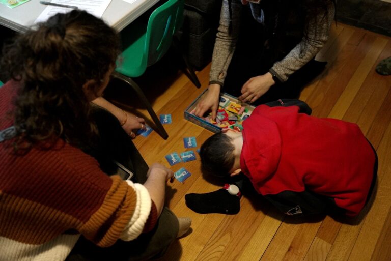 A boy in a red sweater is doubled over face-down in front of a box containing cards. Two women sit on the floor on either side of him.