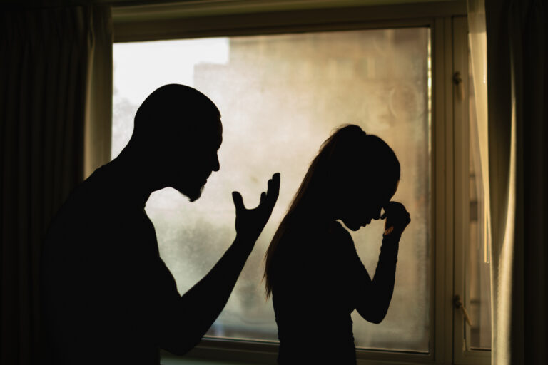 A man gesticulates talking to a woman, who is holding the bridge of her nose in a pose of stress.