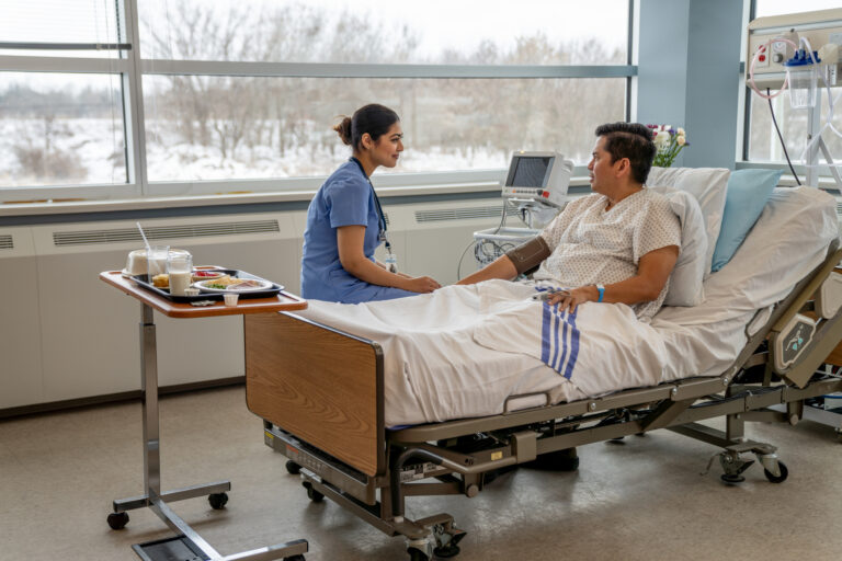 A nurse sits on the edge of a bed talking to a patient. Half eaten food sits on the table nearby.