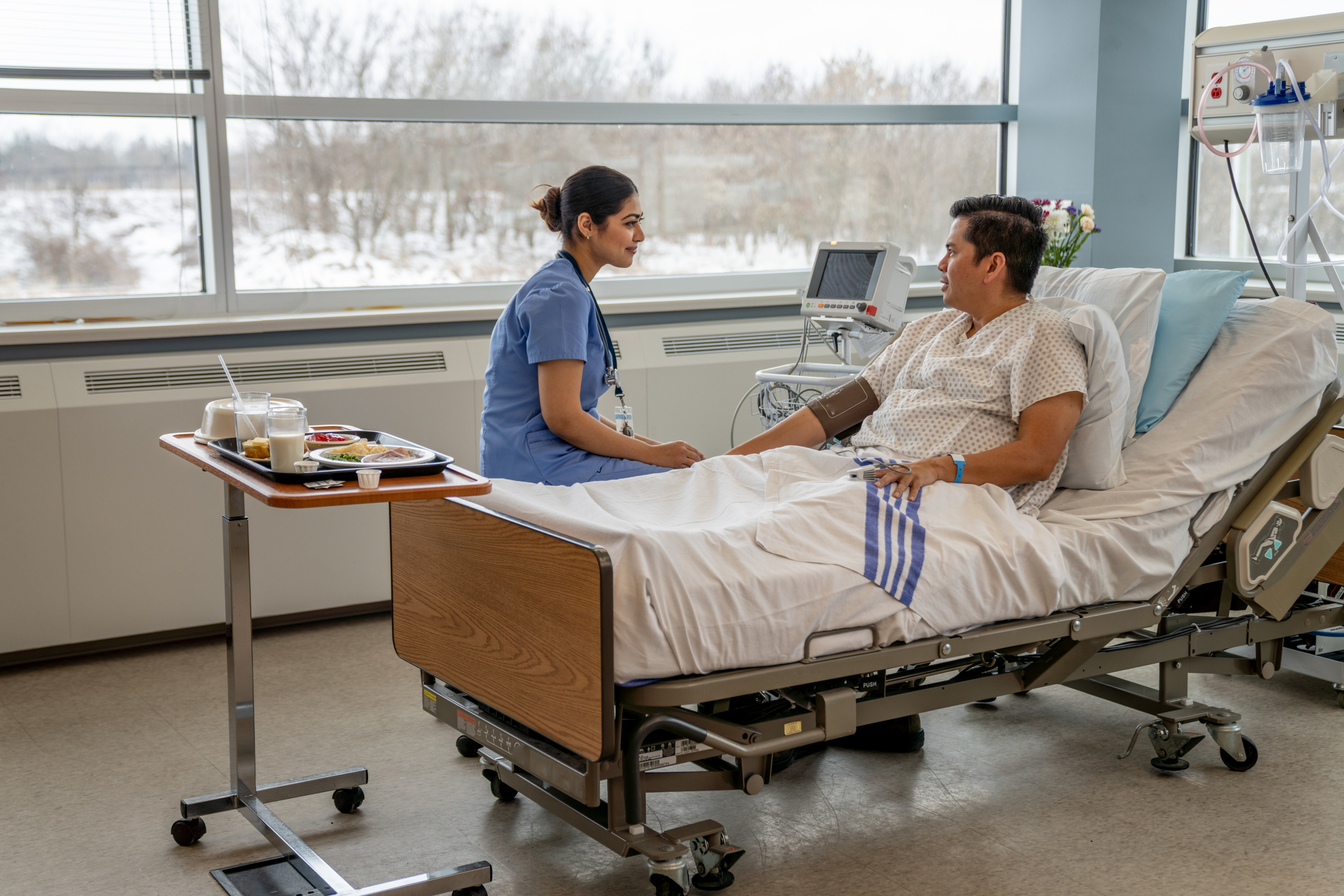 A nurse sits on the edge of a bed talking to a patient. Half eaten food sits on the table nearby.