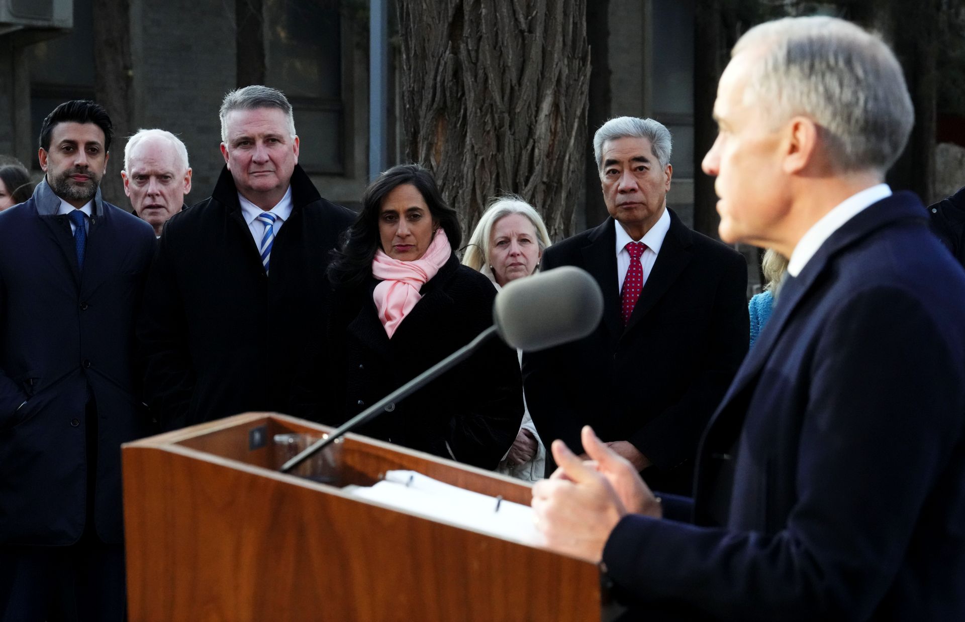 A half-dozen ministers and advisers stand in a line as Prime Minister Mark Carney speaks at a podium. He is in soft focus and in profile while the others are in focus.