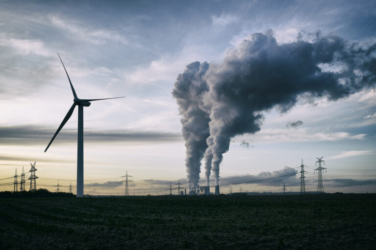 A wind turbine next to a coal-fired power plant with smokestacks belching smoke and power lines in the background.