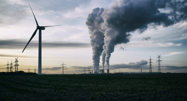 A wind turbine next to a coal-fired power plant with smokestacks belching smoke and power lines in the background.