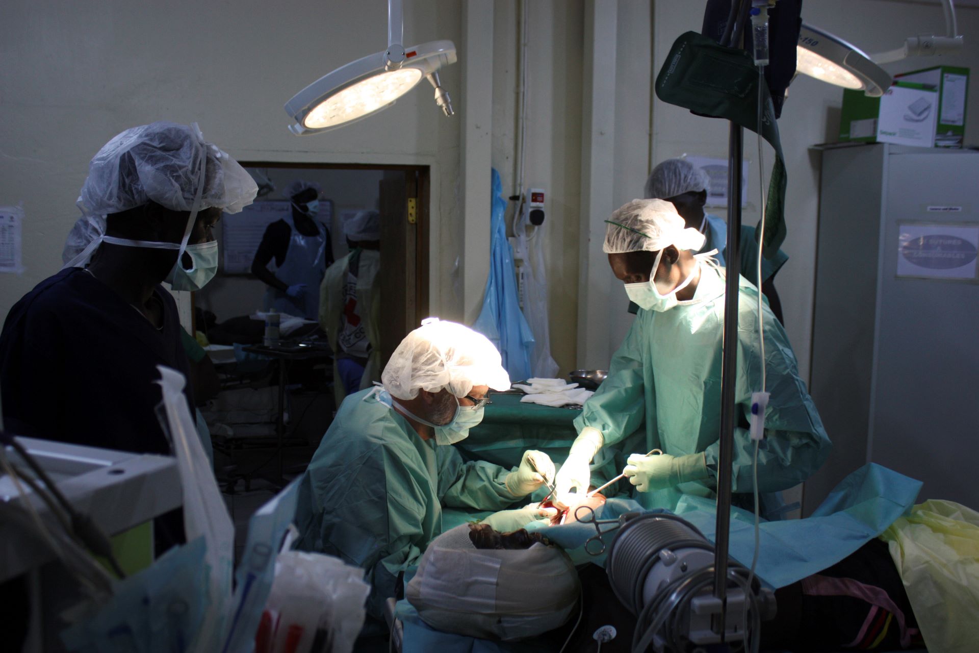 Three doctors operate in a room with two hanging lights and very little else.