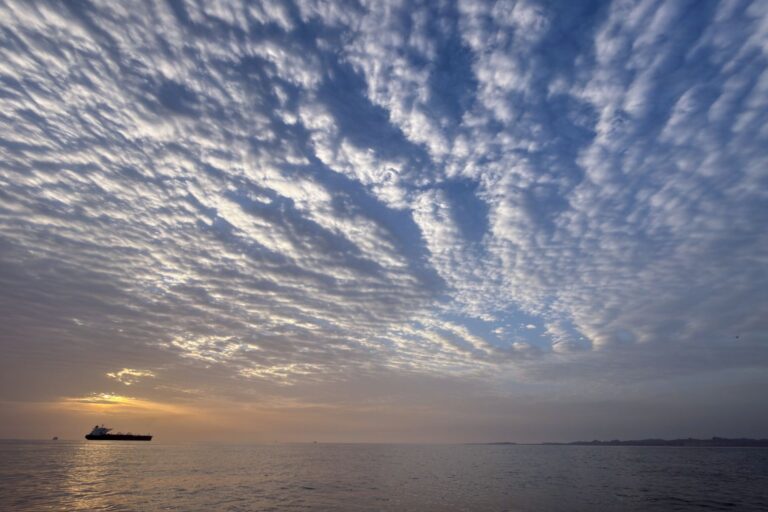The sun rises behind a tanker anchored at sea below the ripples of a partially cloudy sky.