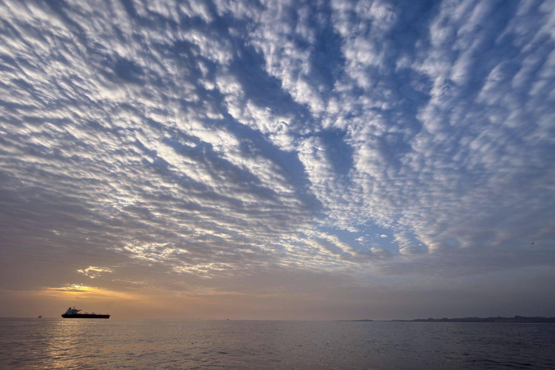 The sun rises behind a tanker anchored at sea below the ripples of a partially cloudy sky.