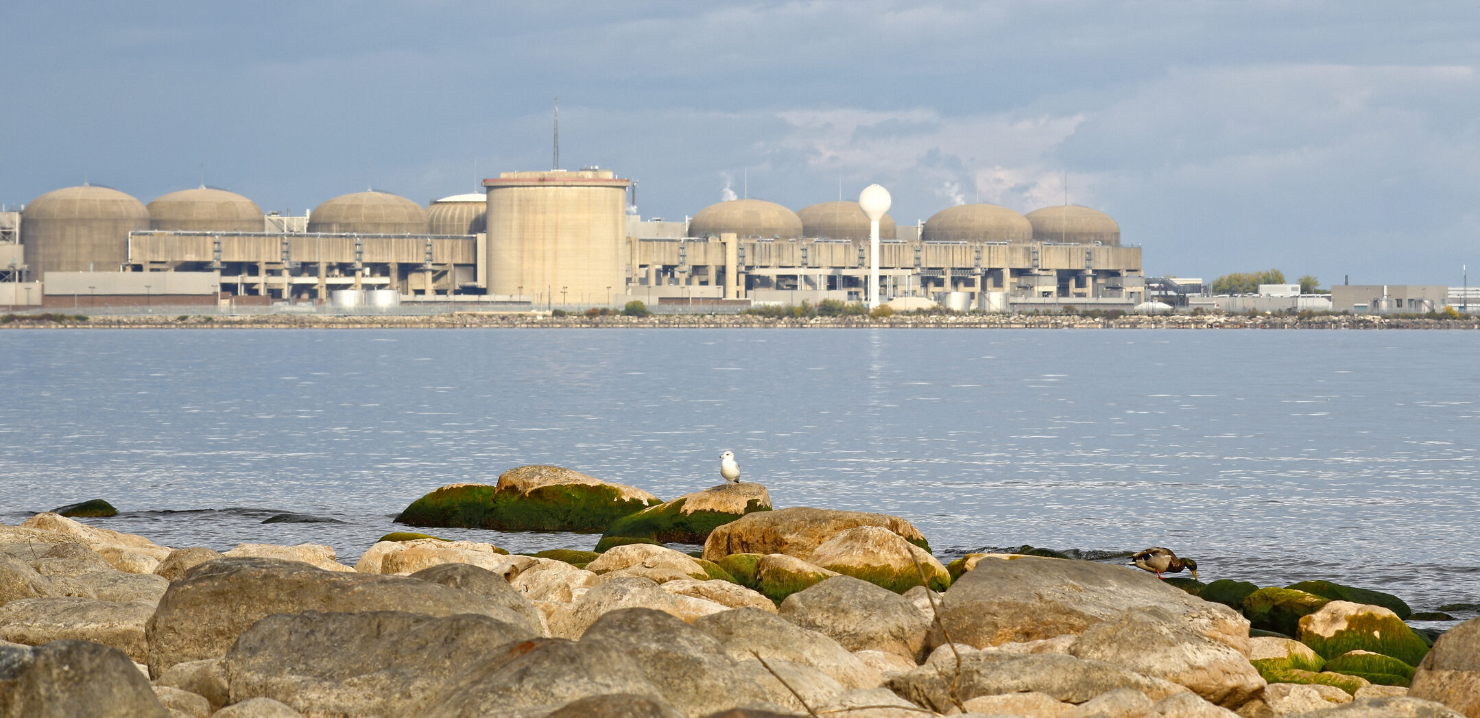 The Pickering Nuclear Generating Station stretches along the shore of Lake Ontario, with several dome-shaped reactor buildings under a cloudy sky, viewed from a rocky foreground by the water.