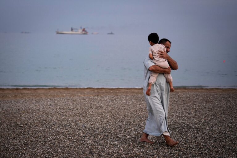 A man carries a child on the beach with oil tankers and cargo ships in the Strait of Hormuz in the background.