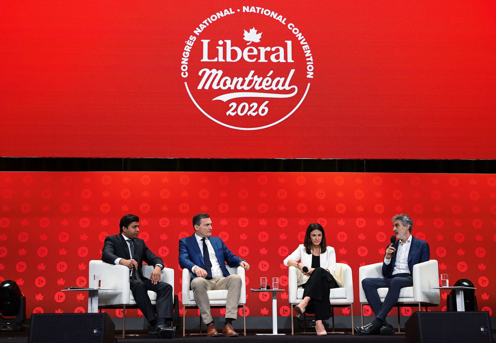Four people sitting on easy chairs holding microphones on a stage in front of Liberal Party of Canada colours and a logo for their 2026 convention.