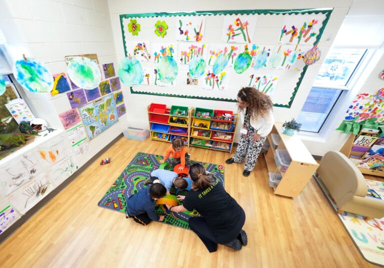 Three children and an adult play a game on a carpet in a child-care centre while another adult looks on.