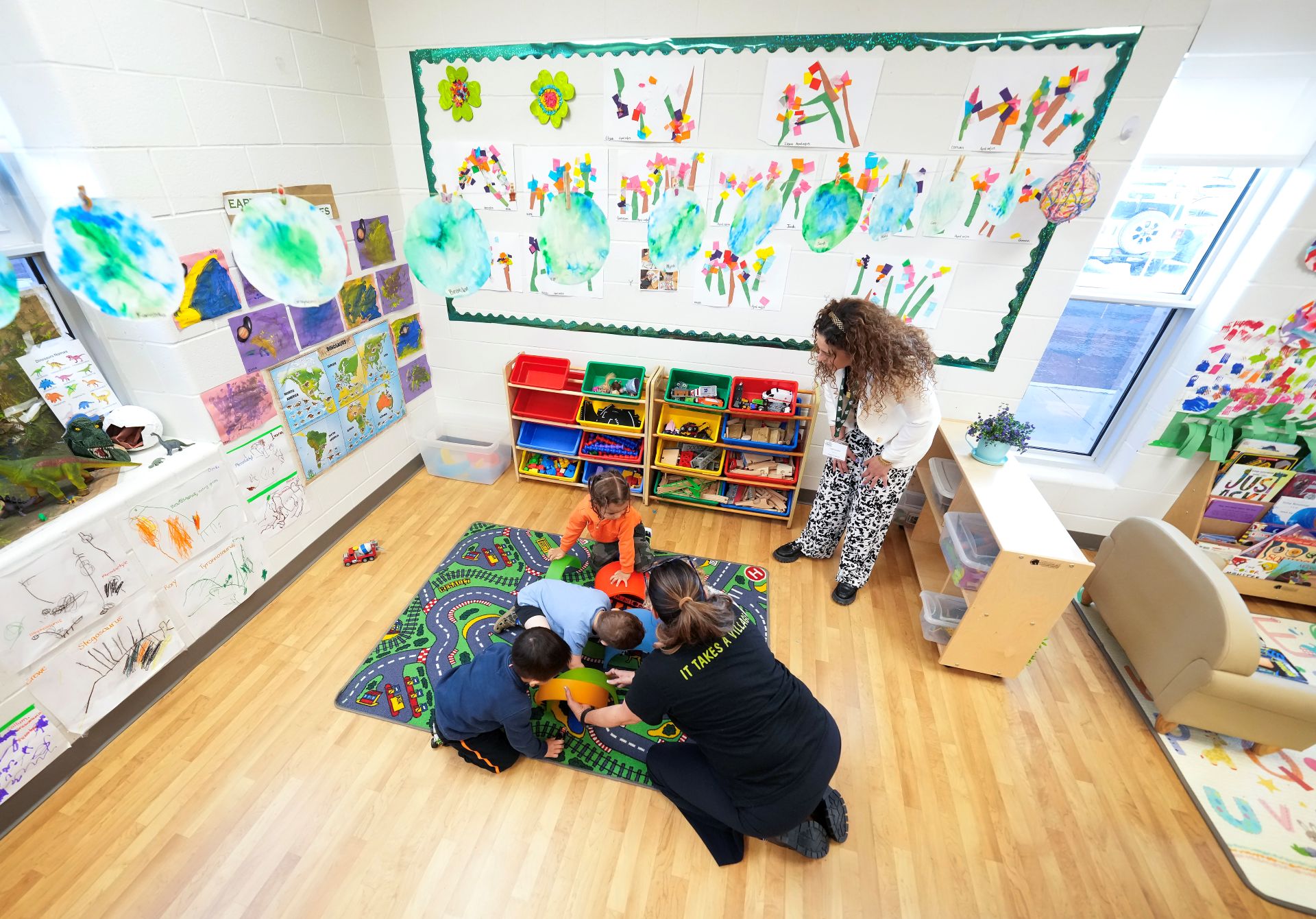 Three children and an adult play a game on a carpet in a child-care centre while another adult looks on.